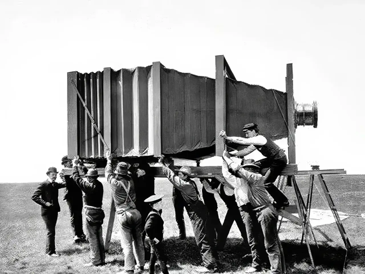 Grupa mężczyzn w historycznych strojach montuje gigantyczny aparat fotograficzny, jakby właśnie wtedy, kiedy wynaleziono aparat fotograficzny.