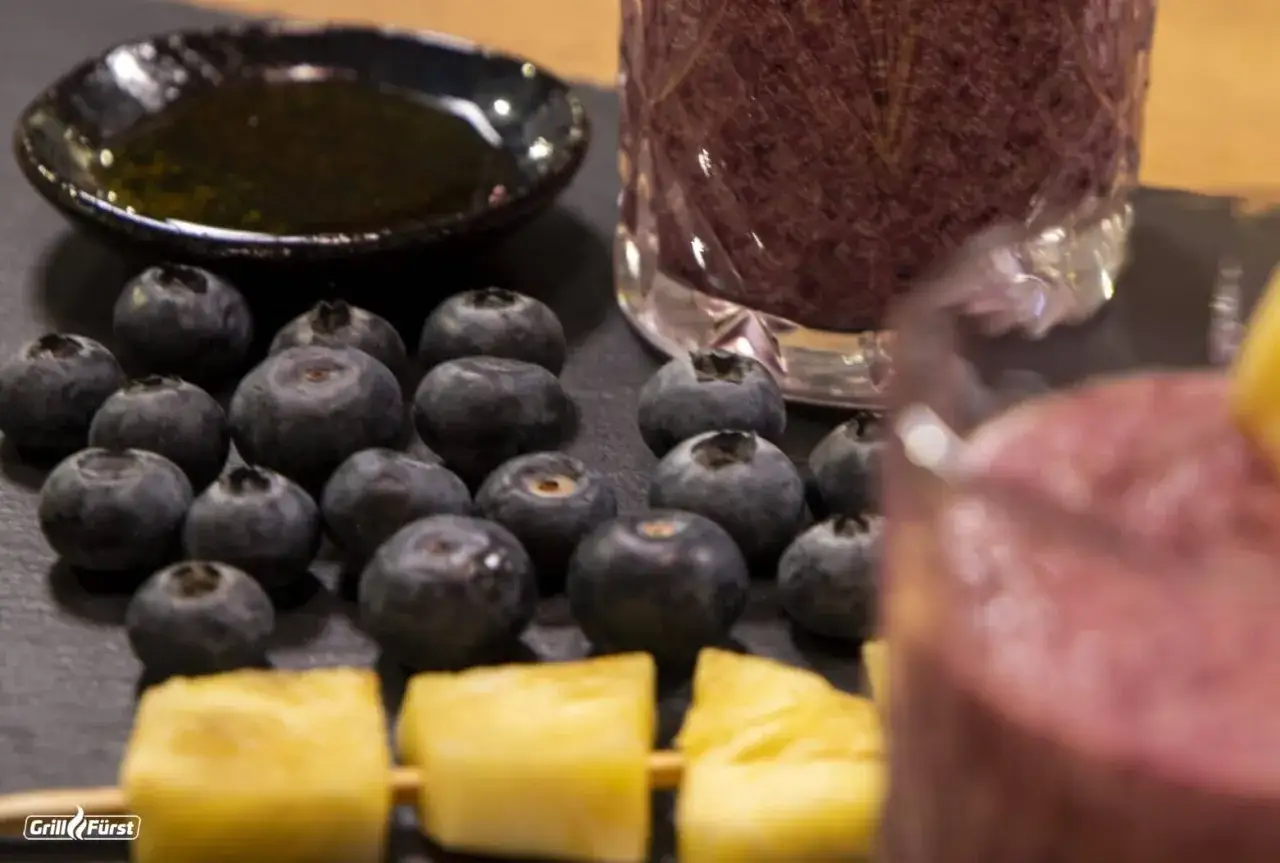 Frische Heidelbeeren warten darauf, verarbeitet zu werden. Daneben ein Glas mit einem lila Smoothie und Ananasstücke auf einem Spieß.