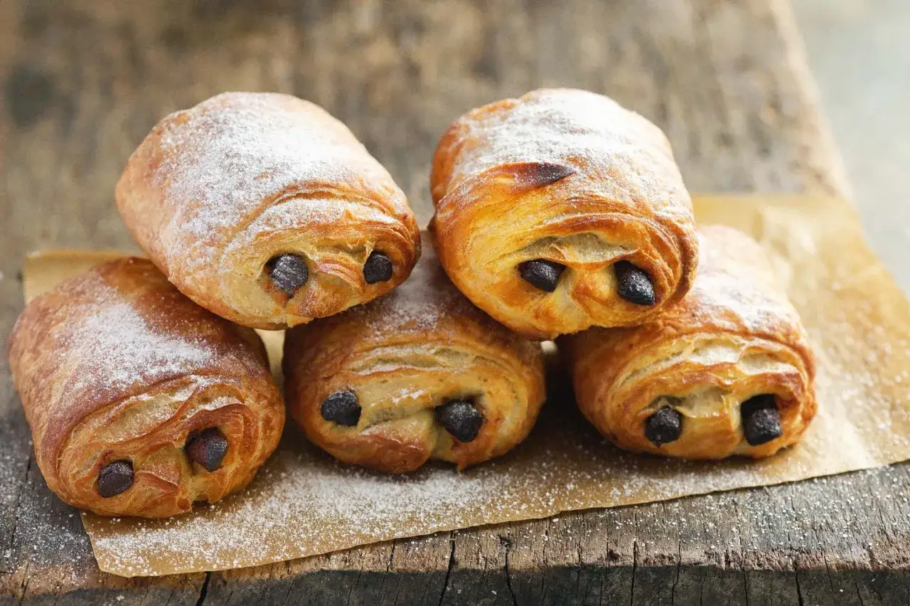 Fünf goldbraune, mit Puderzucker bestäubte Pain au Chocolat liegen auf Backpapier auf einer Holzoberfläche.