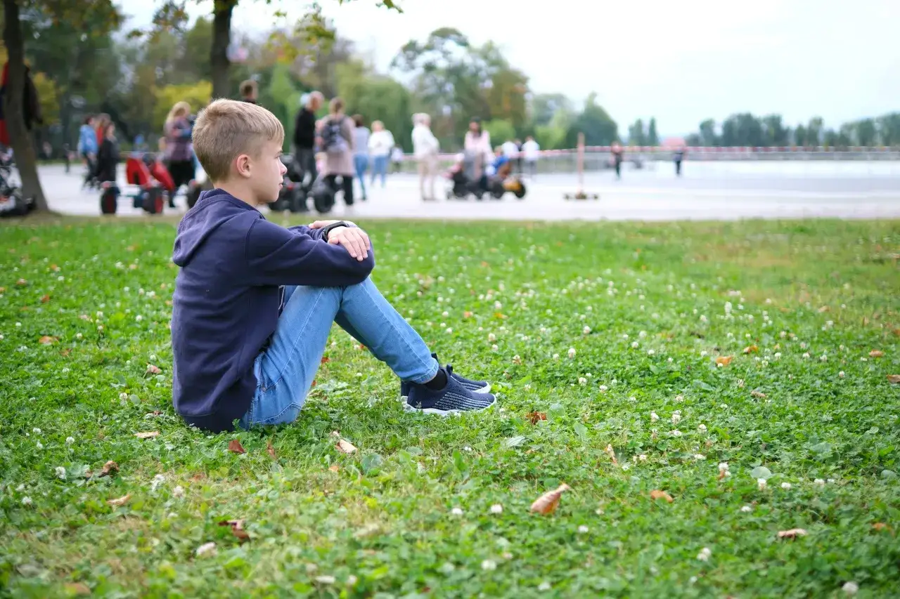 Chłopiec siedzi sam na trawie, otoczony ludźmi. Czy to jedna z przyczyn samotności?