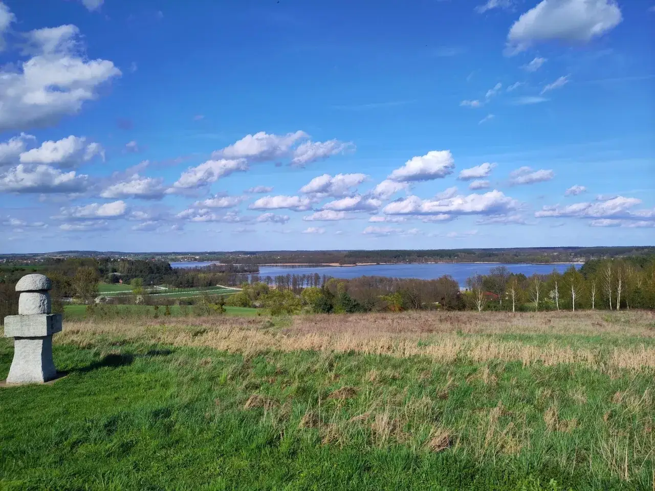 Kamienny obelisk na zielonym wzgórzu, w tle rozległe jezioro Pniewy i błękitne niebo z chmurami.