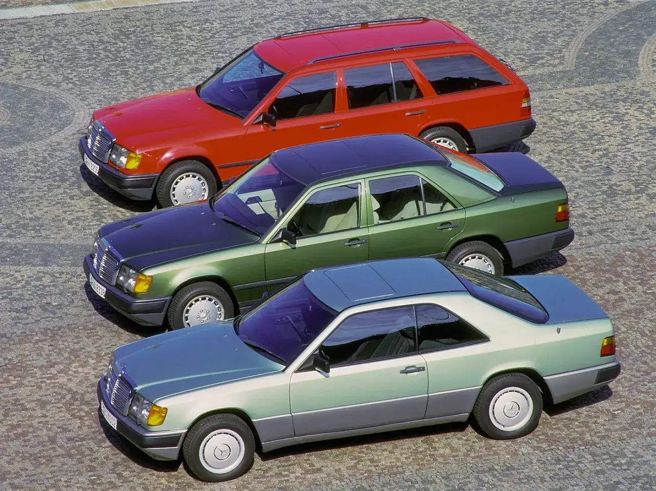 Trzy klasyczne modele w124 Mercedes: czerwone kombi, zielony sedan i zielono-szary coupe, stojące obok siebie na brukowanej nawierzchni.