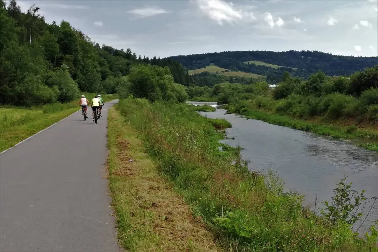 Trzy osoby na rowerach jadą ścieżką rowerową wzdłuż rzeki. Piękny krajobraz Beskidu Sądeckiego zachęca do aktywnego wypoczynku, idealny na velo krynica.