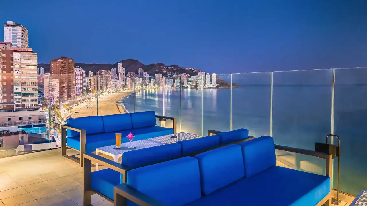 Sofás azules en terraza con vistas a la playa y la ciudad iluminada de Barceló Benidorm Beach al anochecer.