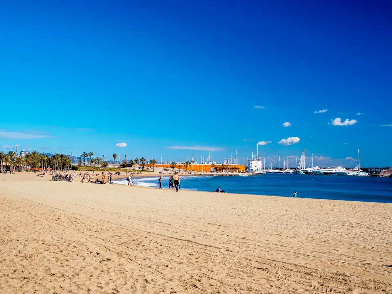 Plaża w Barcelonie, z ludźmi odpoczywającymi na piasku i łodziami w oddali. Czy Barcelona jest nad morzem? Tak, to widać!
