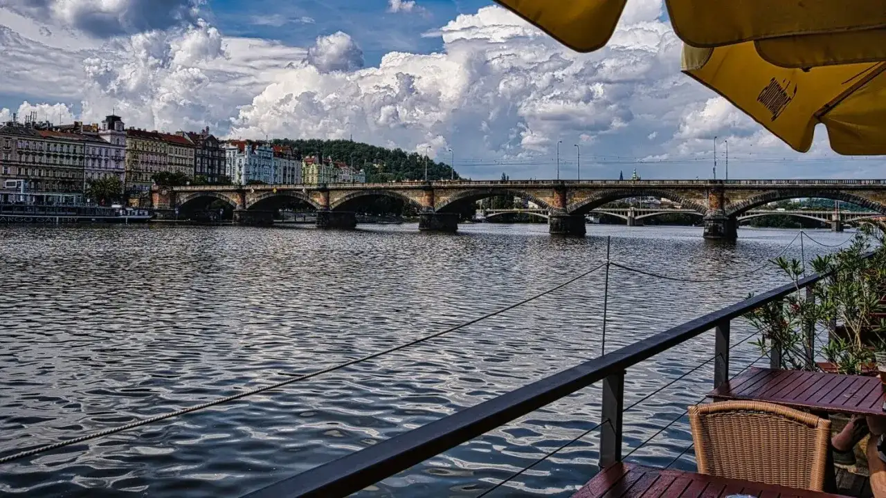 Blick auf die Moldau mit Brücken und Prager Altstadt. Ein Tisch mit Stuhl unter einem Sonnenschirm lädt zum Verweilen ein – ein echter Geheimtipp in Prag.
