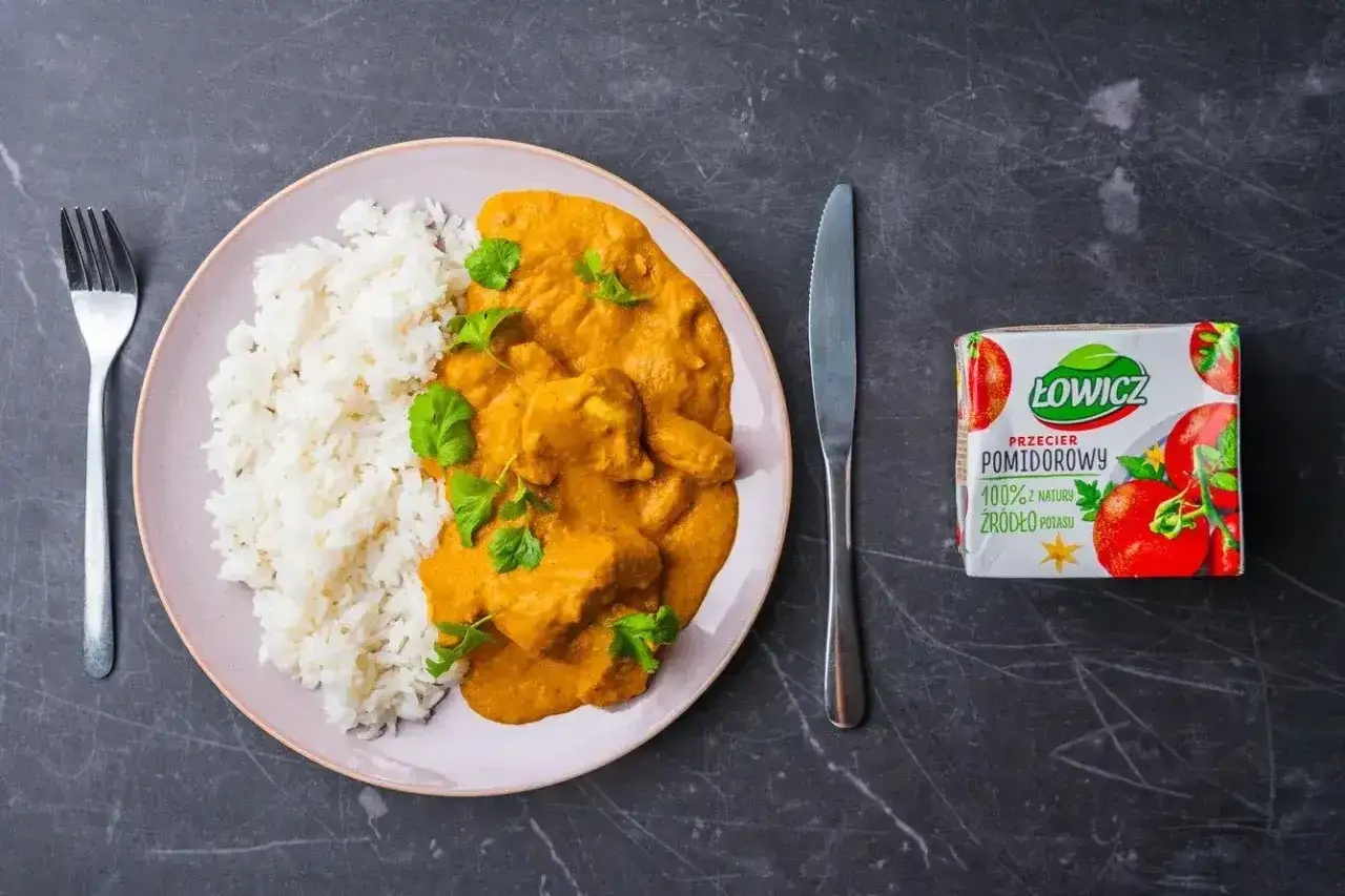 Pyszny butter chicken przepis na talerzu z ryżem, udekorowany świeżą kolendrą. Obok nóż, widelec i przecier pomidorowy Łowicz.
