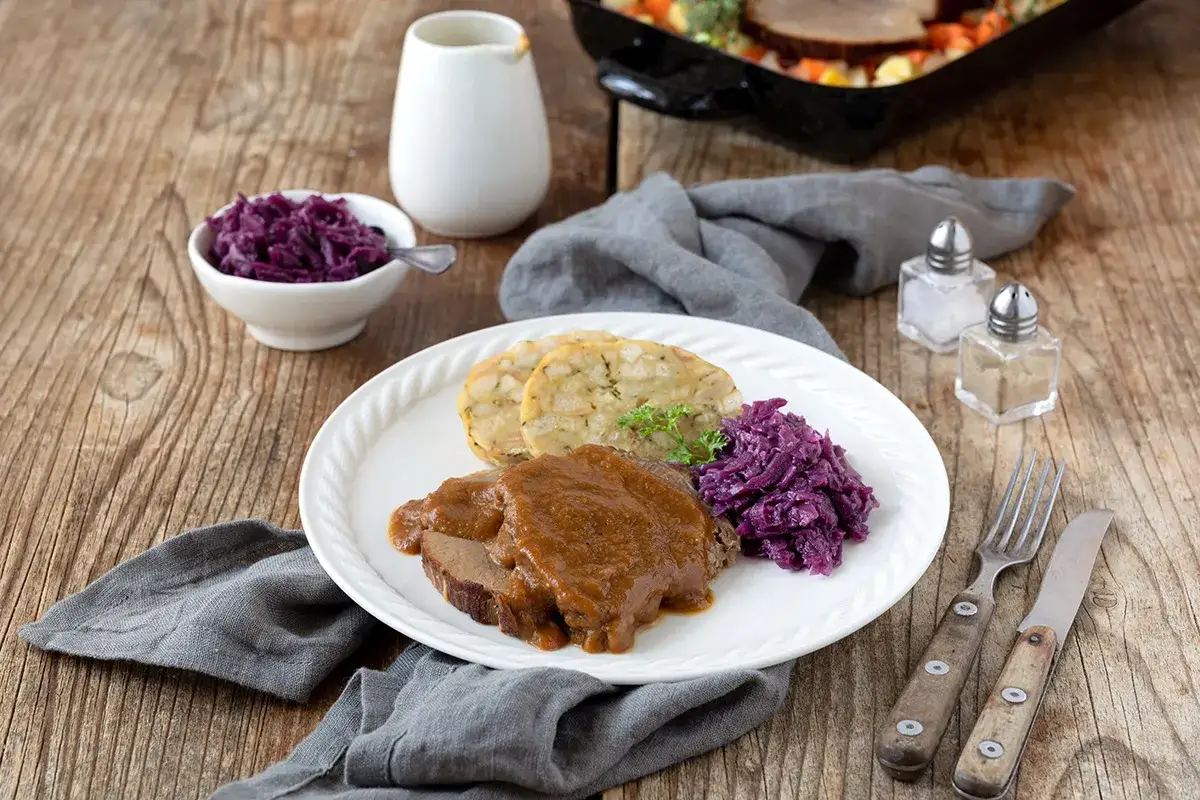 Rinderbraten aus dem Dutch Oven, serviert mit Klößen und Rotkohl auf einem Holztisch.
