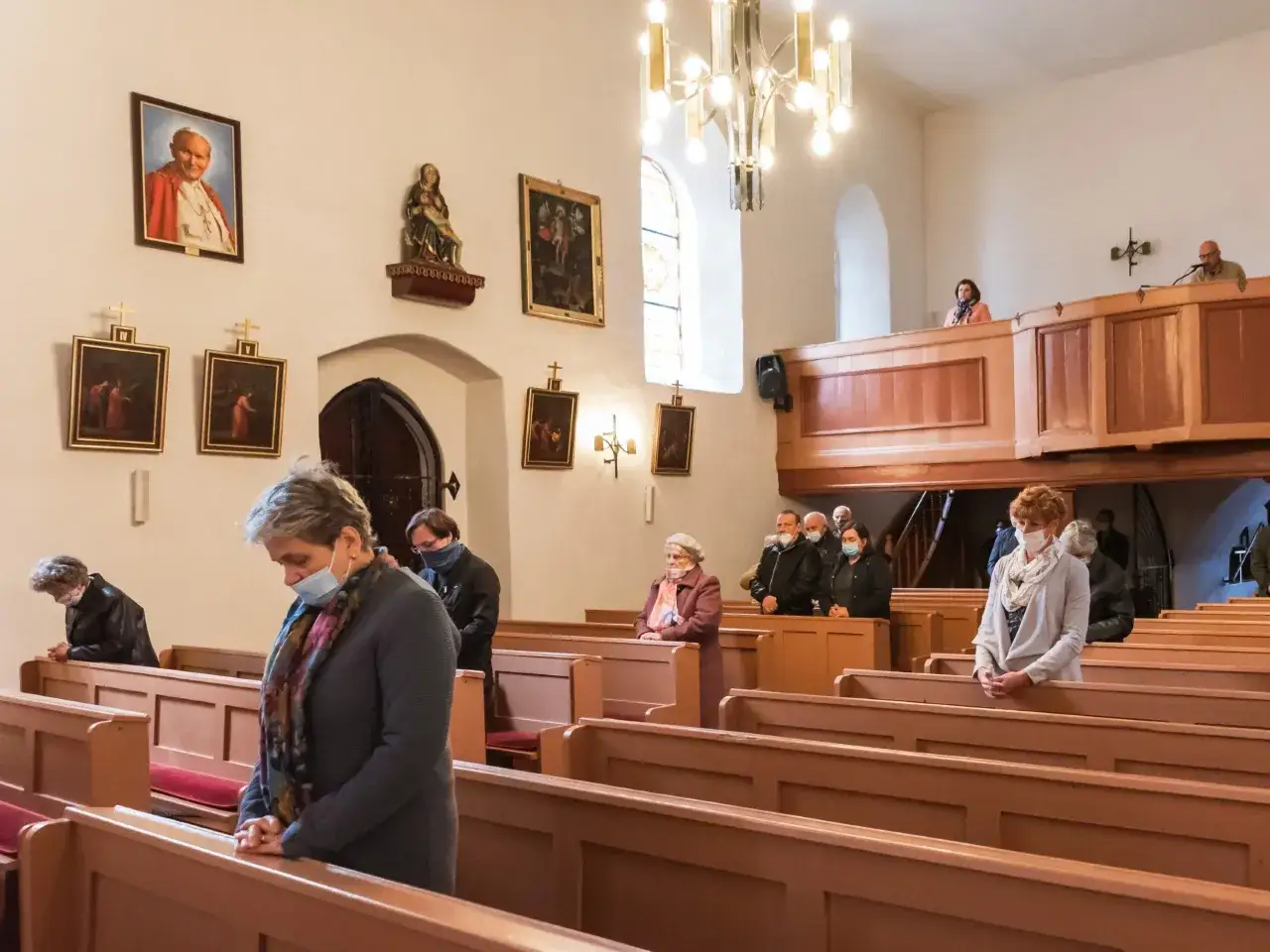 Ludzie w kościele modlą się, zastanawiając się, ile trwa msza święta. Widać portret Jana Pawła II i obrazy religijne.