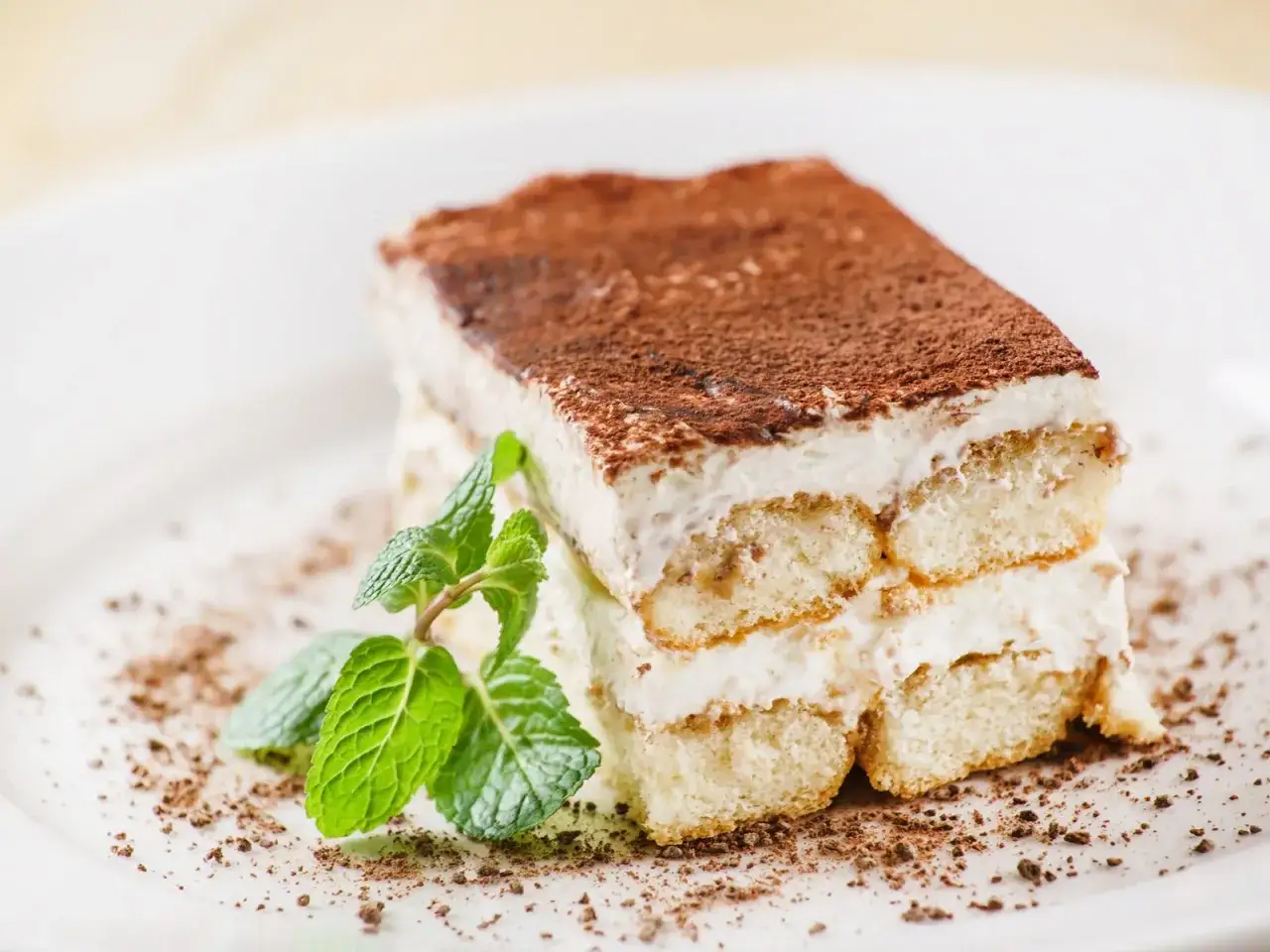 Ein St&uuml;ck original italienisches Tiramisu, best&auml;ubt mit Kakao, garniert mit frischer Minze auf einem wei&szlig;en Teller.