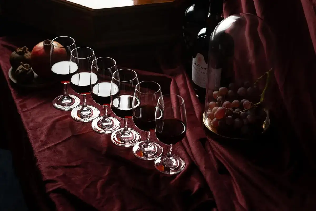 Sechs Gläser mit dunklem Wein, bereit für Cocktails mit Portwein. Daneben Trauben unter einer Glasglocke und eine Flasche Portwein.