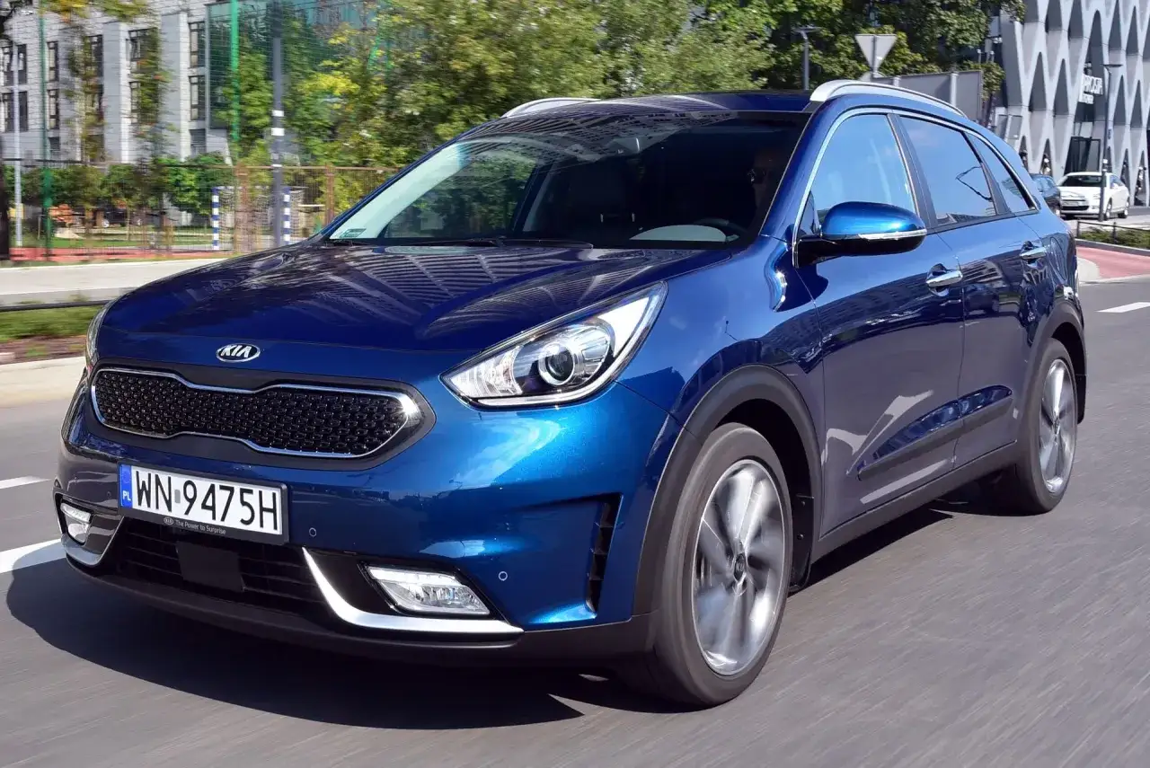 Błękitne Kia Niro jedzie ulicą, odbijając budynki i drzewa.