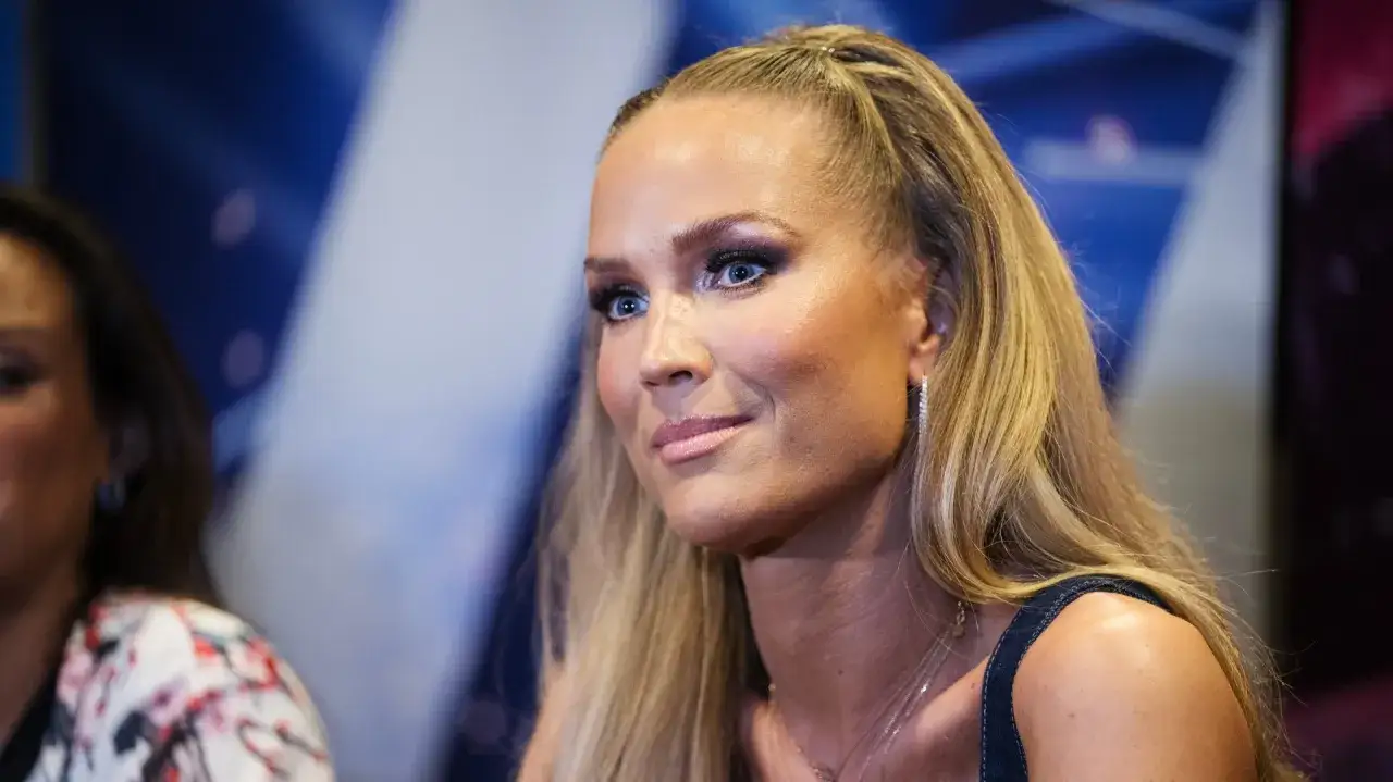 Marie Serneholt med blå ögon och blont hår i en hästsvans, bär smycken och ser åt sidan.