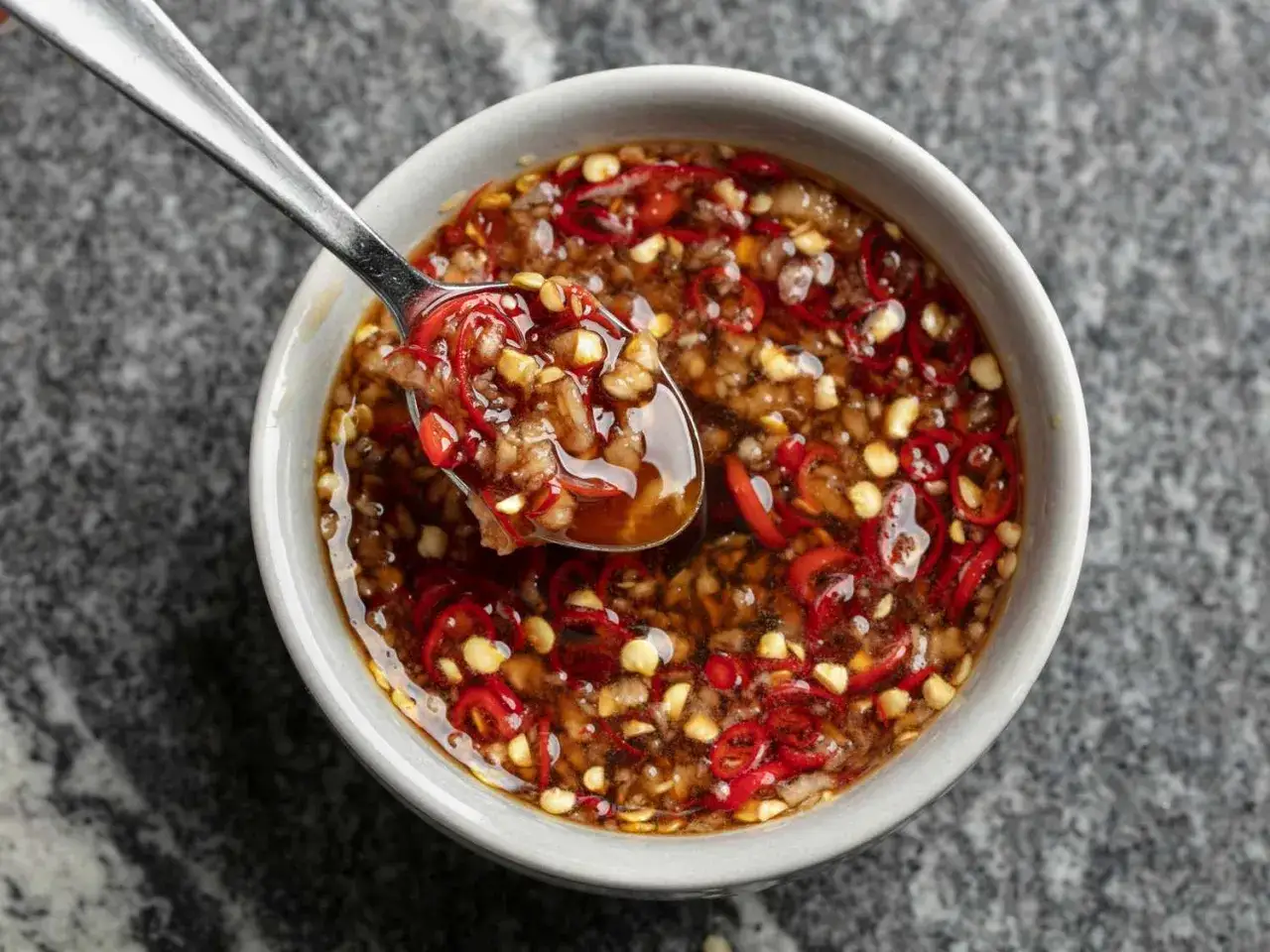 Eine Schüssel mit scharfer Thai Chilli Sauce, auf einem Löffel sind rote Chilischeiben und Knoblauch zu sehen.