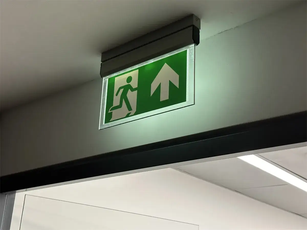 Przegląd oświetlenia awaryjnego i ewakuacyjnego. Zielony znak z biegnącą postacią i strzałką w górę, zamontowany na suficie.