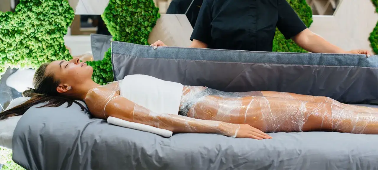 Kobieta leży na łóżku, owinięta folią podczas zabiegu body wrapping. W tle zielone dekoracje.