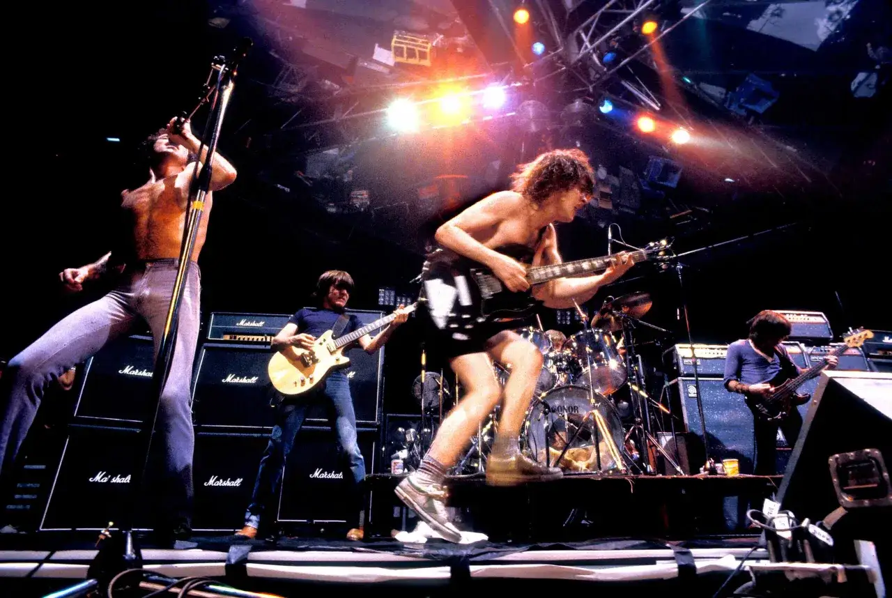 Gitarzysta AC/DC w szaleństwie na scenie, z rozpuszczonymi włosami i w krótkich spodenkach, gra solo.