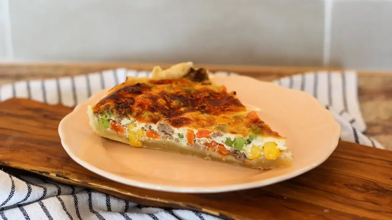 Ein St&uuml;ck herzhafte vegetarische Quiche mit Karotten, Erbsen und Mais auf einem rosa Teller.