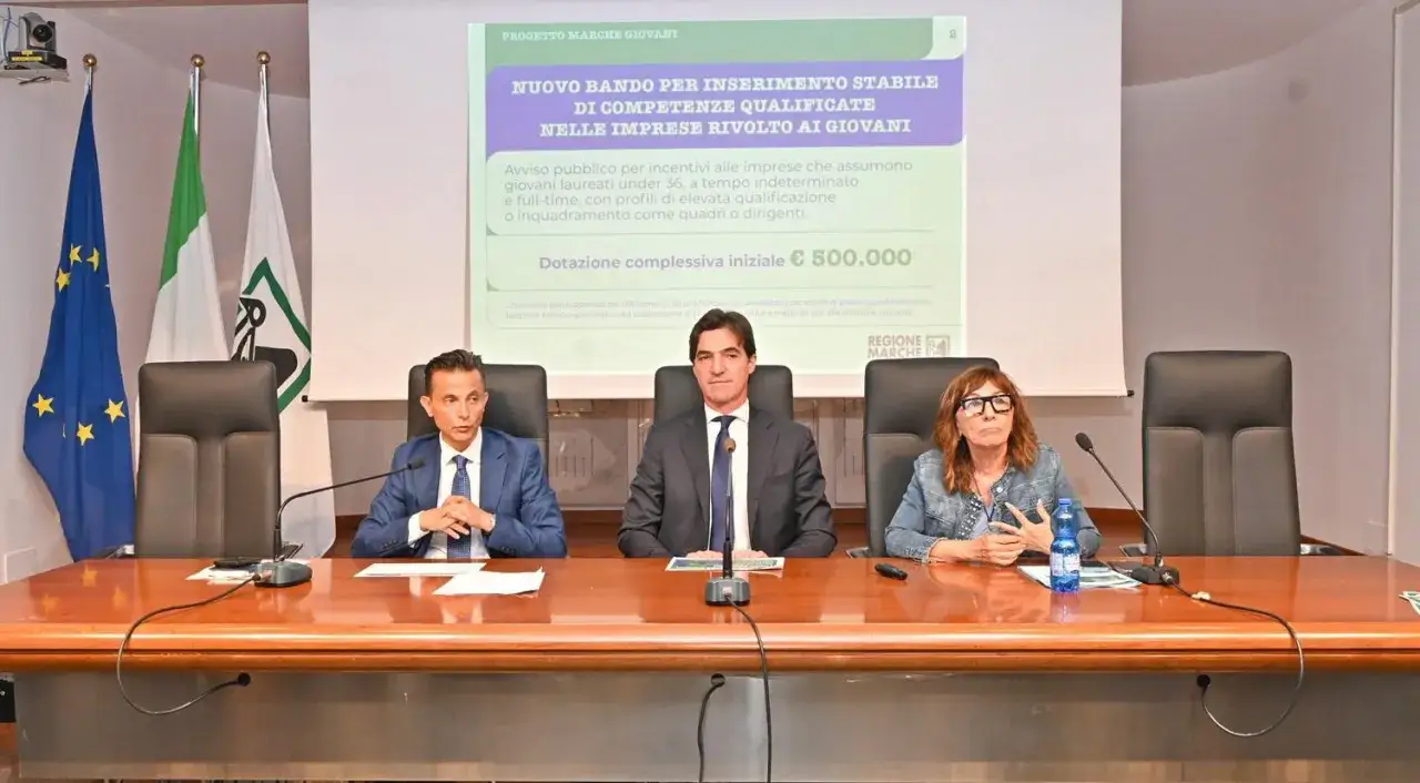 Presentazione di un bando per il **talent acquisition** di giovani qualificati. Tre persone siedono a un tavolo, con bandiere e uno schermo sullo sfondo.