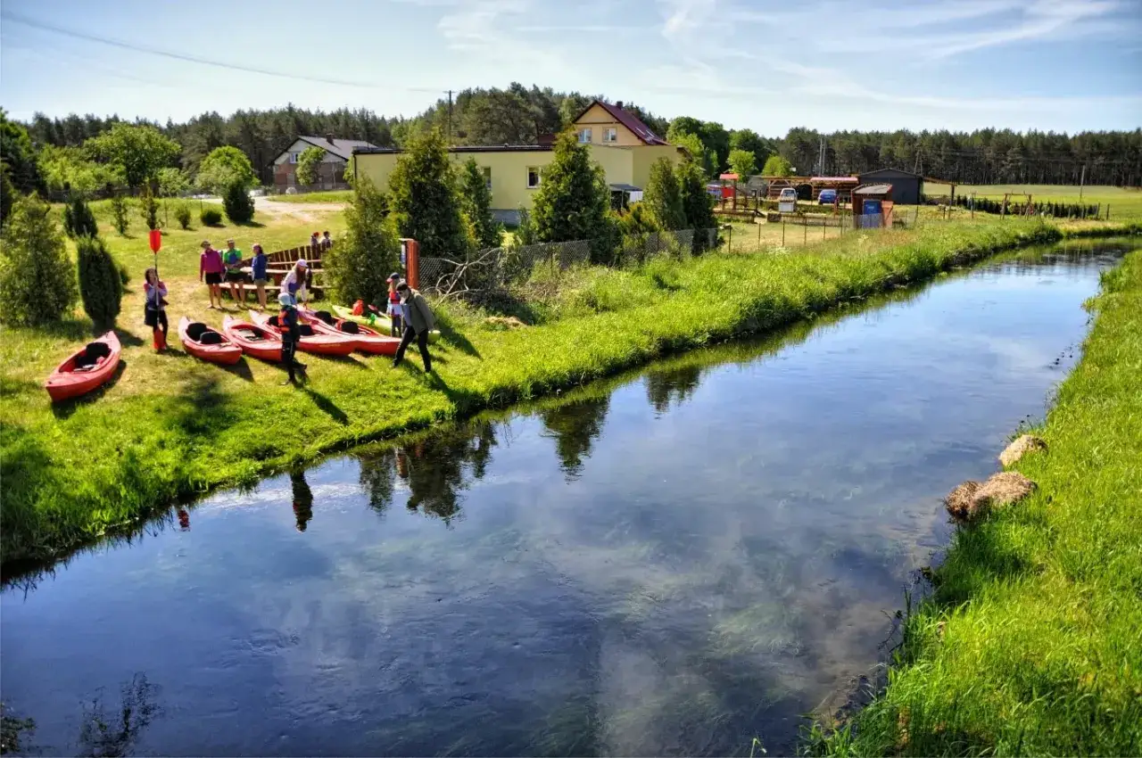 Grupa ludzi przygotowuje się do spływu kajakowego na Kaszubach. Czerwone kajaki czekają na brzegu rzeki.