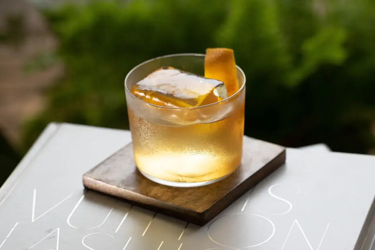 Un verre de cocktail ambré, orné d'un zeste d'orange et d'un gros glaçon transparent, repose sur un sous-verre en bois.