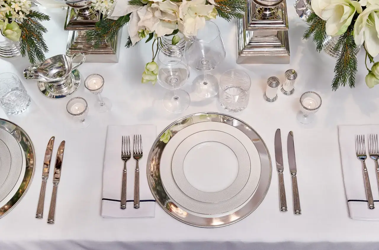 Elegant dukning med silverf&auml;rgat bestick, vita servetter och porslin. Blomsterarrangemang och ljus skapar en festlig atmosf&auml;r.
