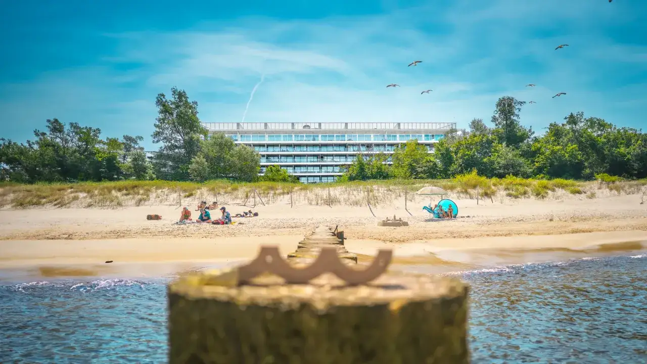 Słoneczne Kołobrzeg wczasy z wyżywieniem. Ludzie odpoczywają na plaży przed nowoczesnym hotelem, mewy szybują na niebie.