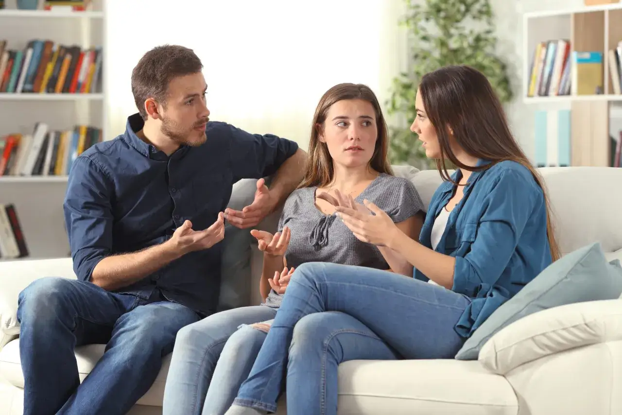 Tres personas sentadas en un sofá blanco discuten animadamente, gesticulando con las manos, en un intento por resolver conflictos.