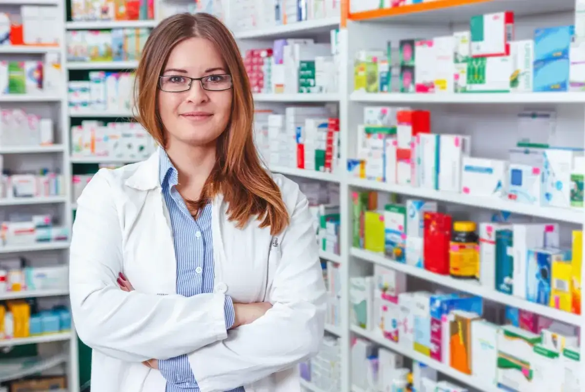 Kobieta w białym fartuchu stoi na tle apteki. Zastanawiasz się, ile zarabia technik farmacji?