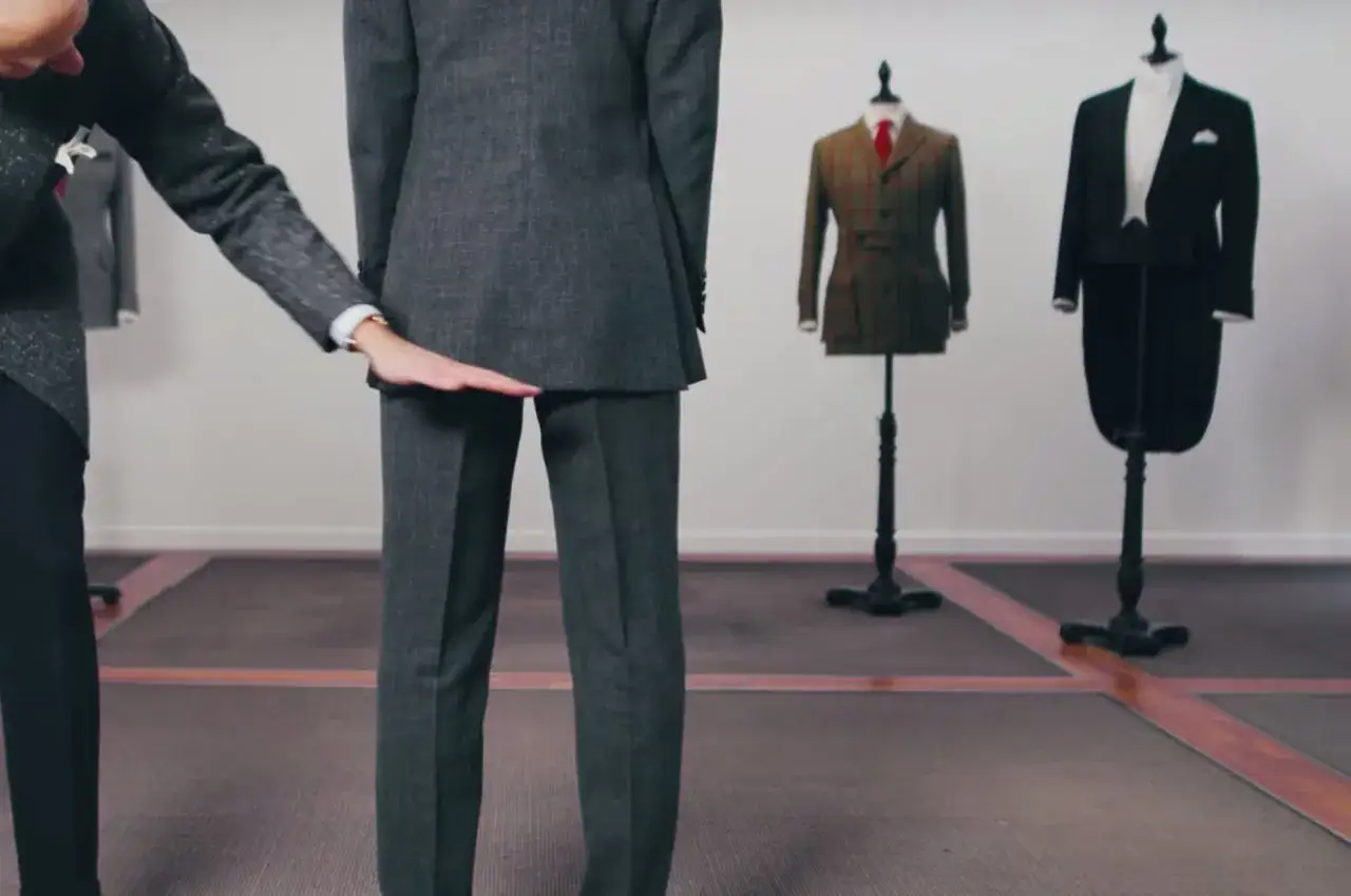 A tailor demonstrates how long should suit jacket be, with mannequins in the background showing different suit styles.