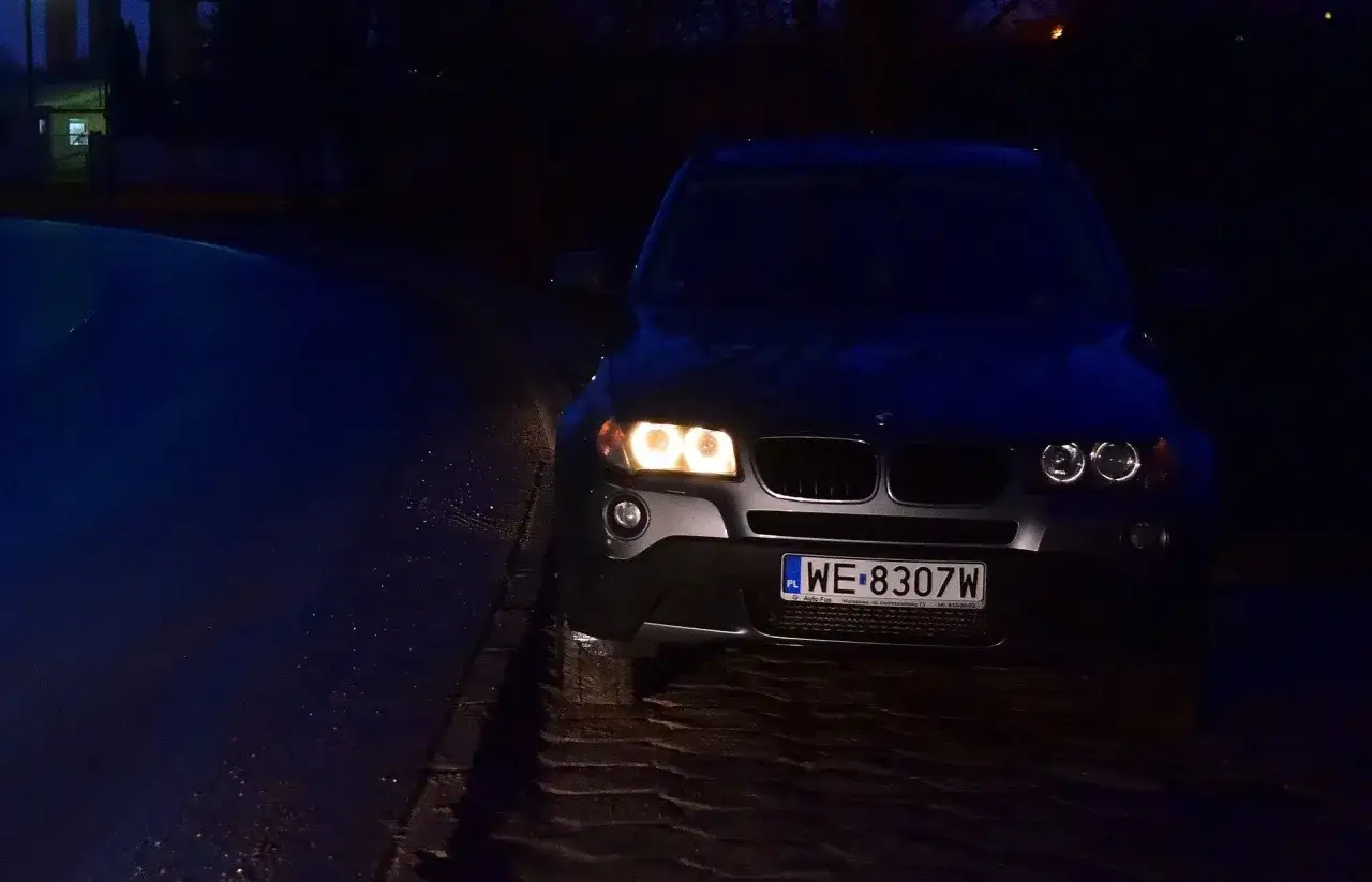 Ciemny samochód z włączonymi światłami postojowymi na mokrej, brukowanej drodze.