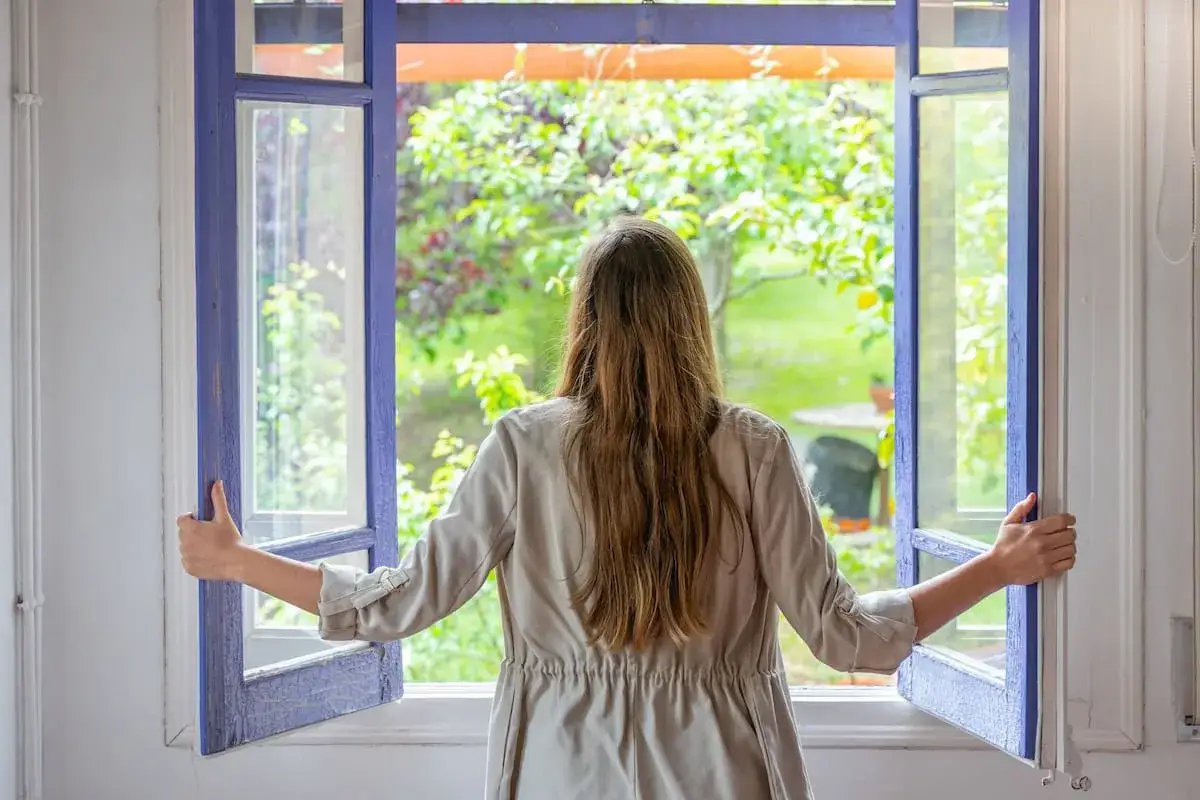 Mujer abriendo ventana para purificar el aire de casa de forma natural. El aire fresco del jardín entra en la habitación.