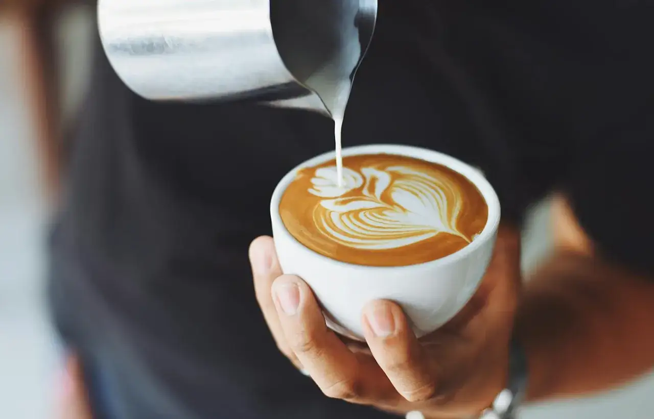 Ręka trzyma filiżankę z idealnym flat white, na którym barista tworzy wzór z mlecznej pianki.