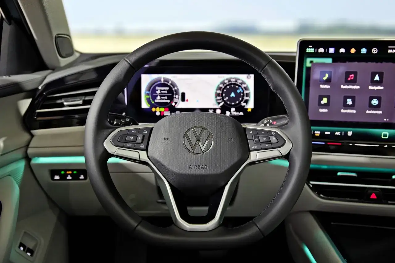 Wnętrze Volkswagena z kierownicą i ekranami. W tle widać krajobraz, jakby podróżował pasatem.