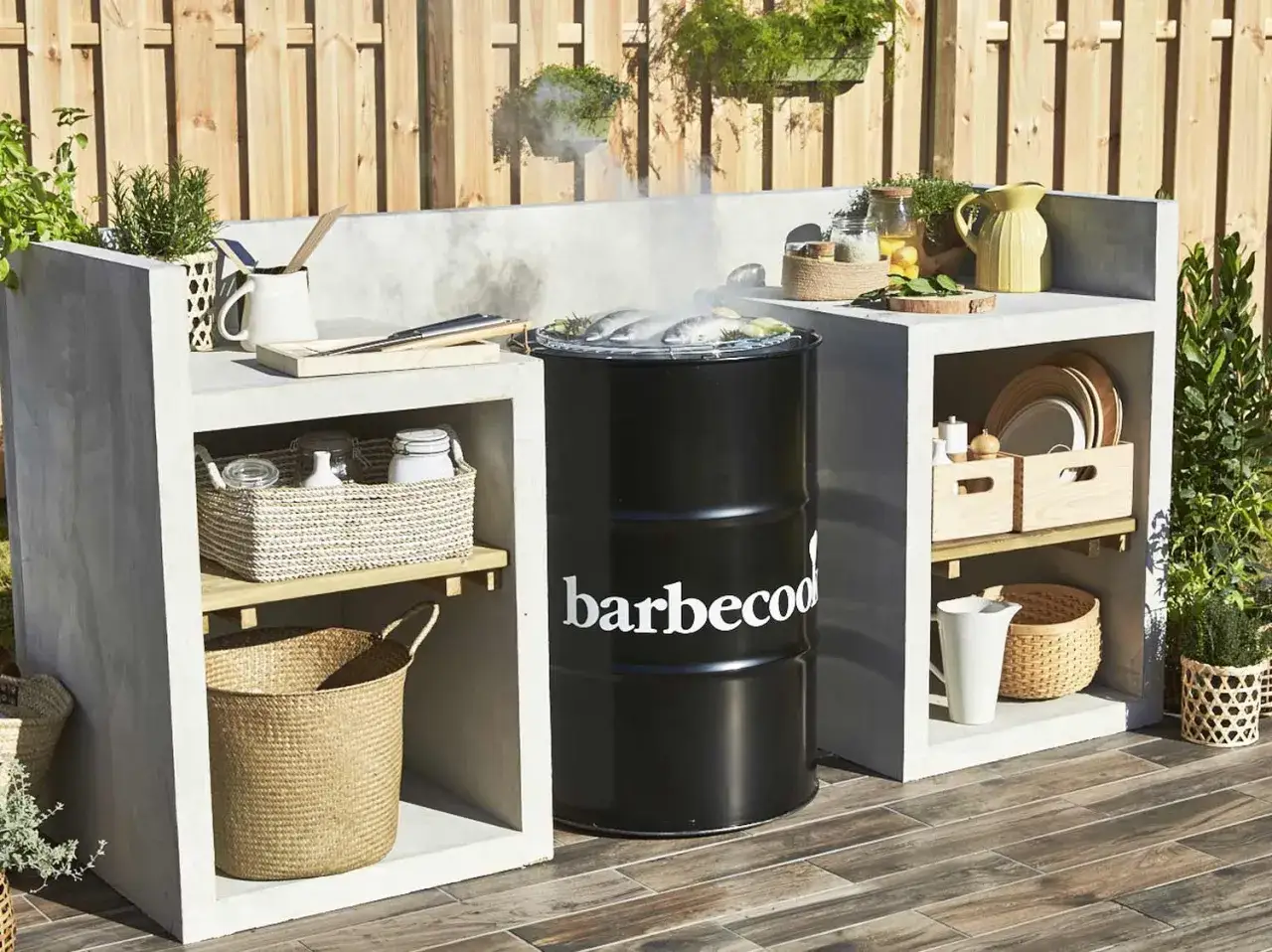 Barbecue d'été fait maison : un fumoir en baril noir avec des poissons sur la grille, intégré dans une cuisine extérieure en béton avec paniers et vaisselle.