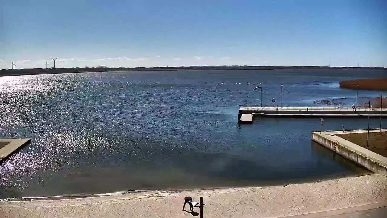 Widok z jeziorsko kamera na żywo. Na piaszczystej plaży dwie osoby, w tle spokojne jezioro, pomost i wiatraki.
