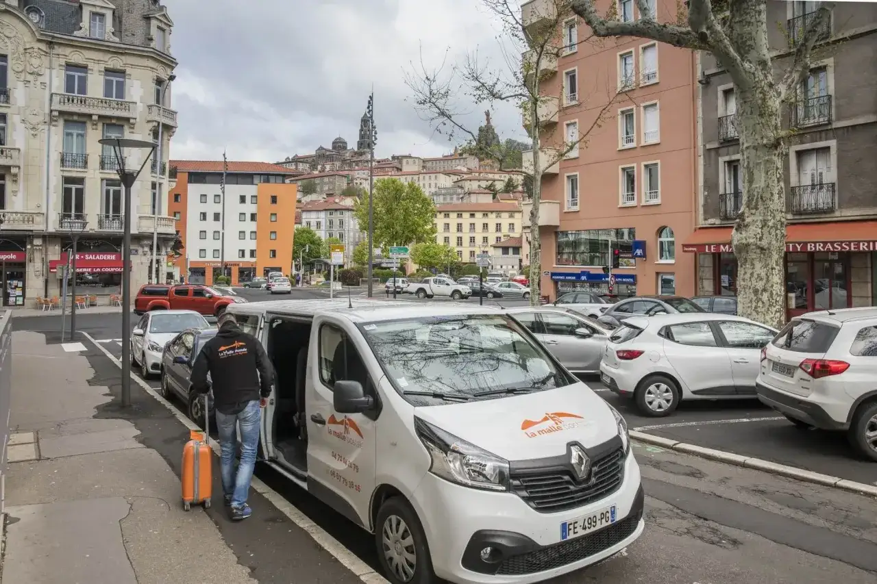 Un homme tire une valise orange vers un bus compostelle blanc. La ville s'étend en arrière-plan.