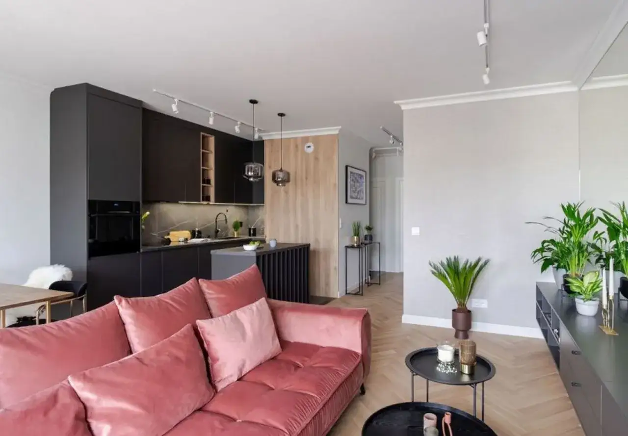 Nowoczesny salon z aneksem kuchennym w projekcie mieszkania 70m2. R&oacute;żowa kanapa, czarne meble kuchenne i drewniane akcenty tworzą przytulną przestrzeń.