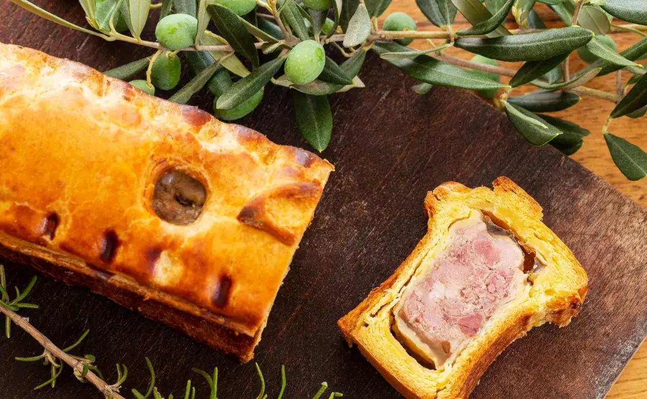 Un plat lorrain app&eacute;tissant, une tranche de p&acirc;t&eacute; en cro&ucirc;te dor&eacute;e et garnie de viande rose, sur fond de bois.