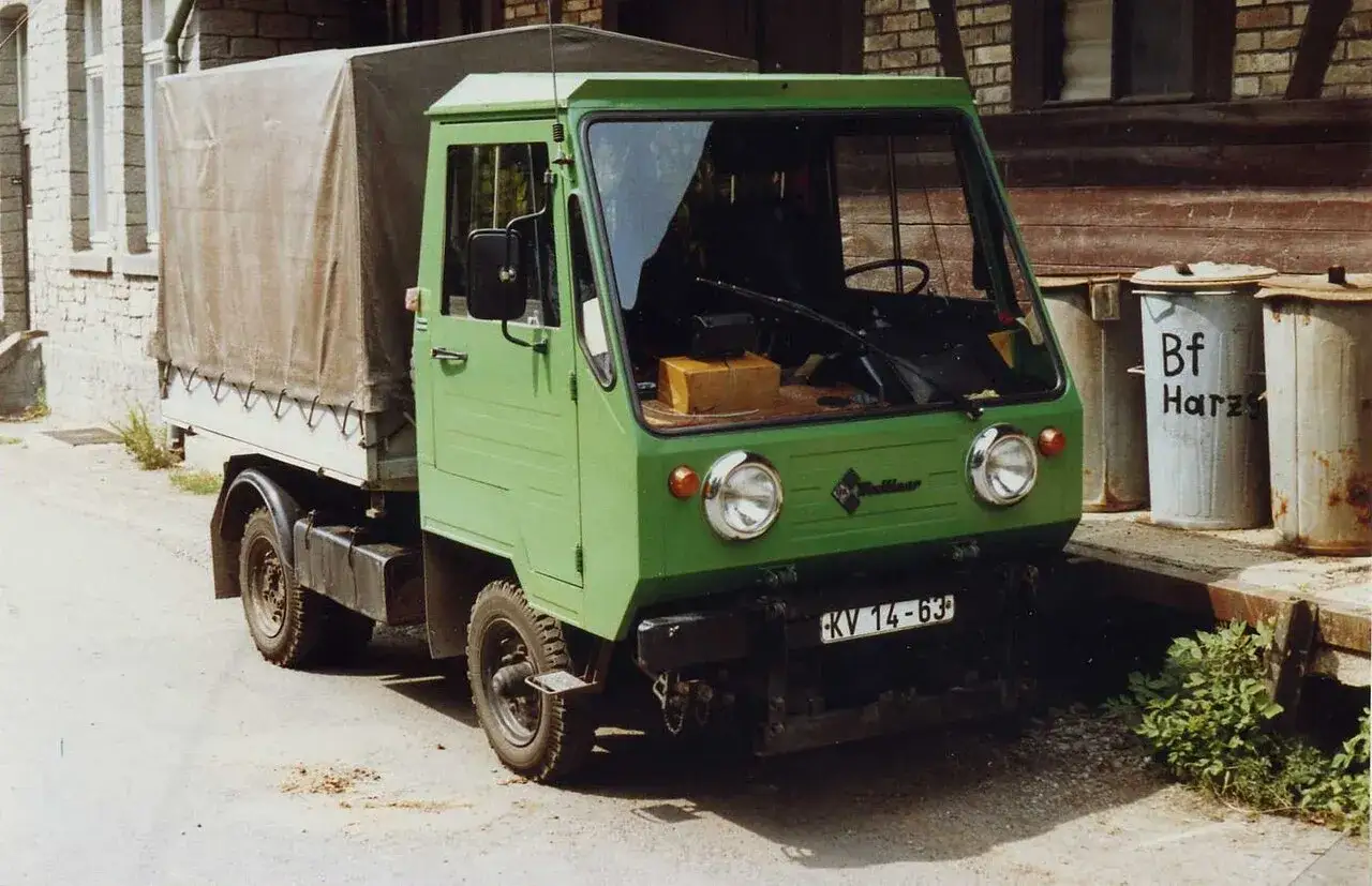 Zielony Multicar M25 z plandeką, gotowy do drogi. Obok śmietniki z napisem 