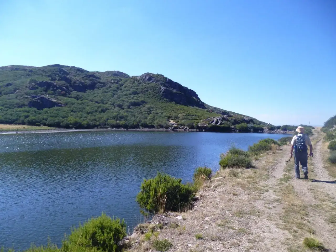 Un excursionista camina por un sendero junto a un lago azul, con montañas verdes de fondo. El aire fresco de xares a veiga invita a la aventura.