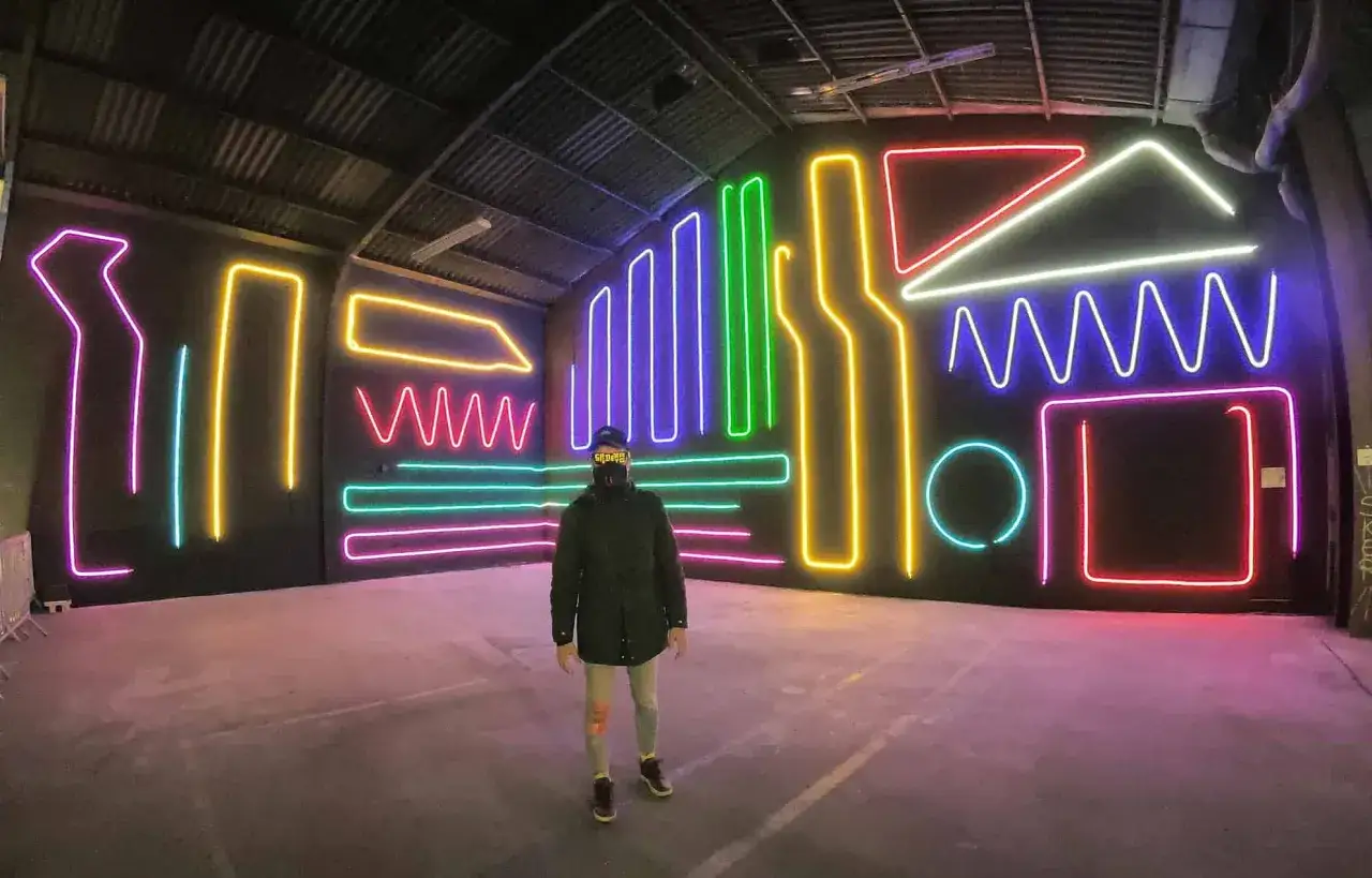 Persona con gorra y bufanda frente a un muro con luces de neón de colores. Inspiración para nombres para cantantes de trap.