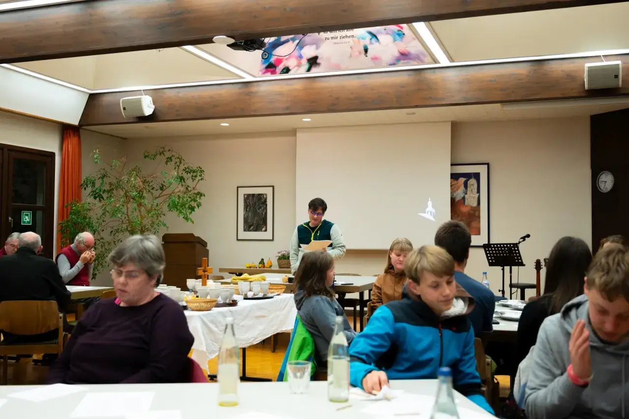 Menschen sitzen an Tischen in einem Raum, der für eine kirchliche Veranstaltung vorbereitet ist. Ein junger Mann steht vorne und hält Dokumente.