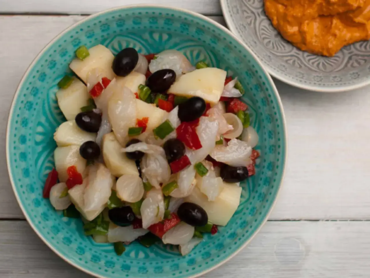 Ensalada de bacalao con patatas, aceitunas negras, pimientos y cebolla morada en un cuenco turquesa.