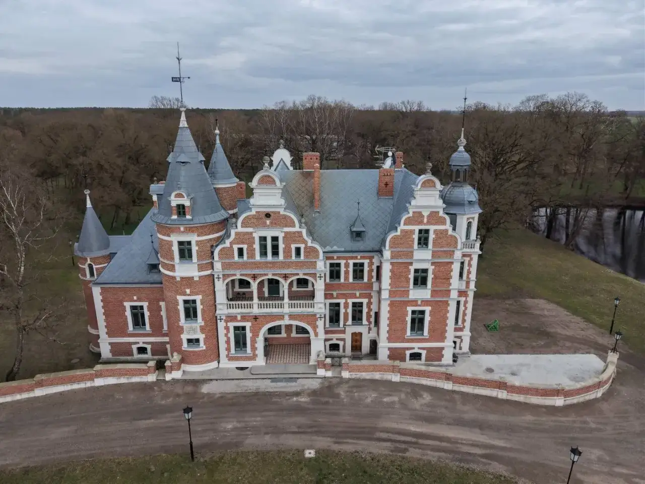 Pałac Kobylniki, z czerwonej cegły, z wieżyczkami i ozdobnym frontem, otoczony drzewami i wodą.