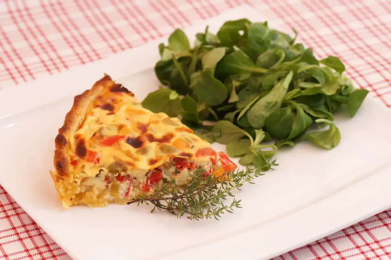Une part de tarte aux poivrons color&eacute;s, garnie d'une branche de thym, accompagn&eacute;e d'une salade verte fra&icirc;che sur une assiette blanche.