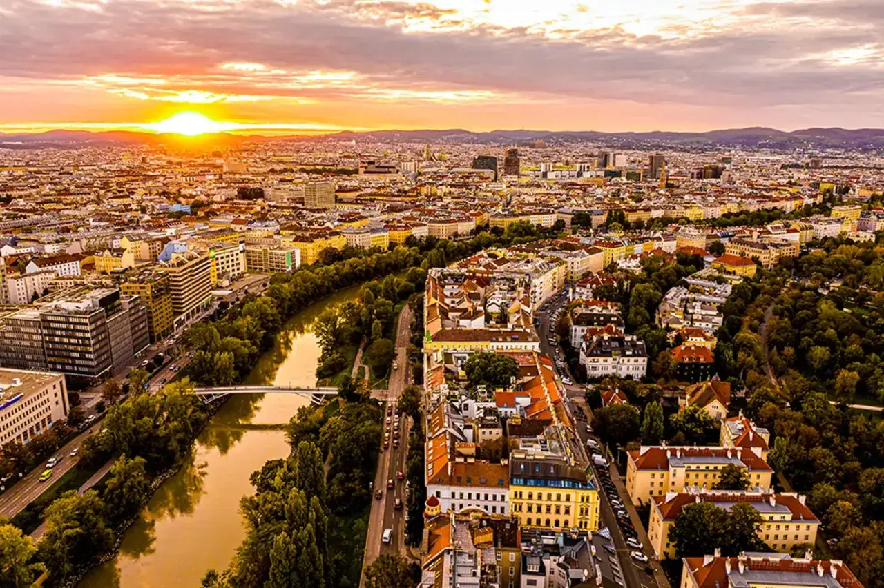 Sonnenuntergang &uuml;ber Wien. Ein Fluss schl&auml;ngelt sich durch die Stadt, umgeben von B&auml;umen und Geb&auml;uden. Ein atemberaubender Blick auf Wien von oben.