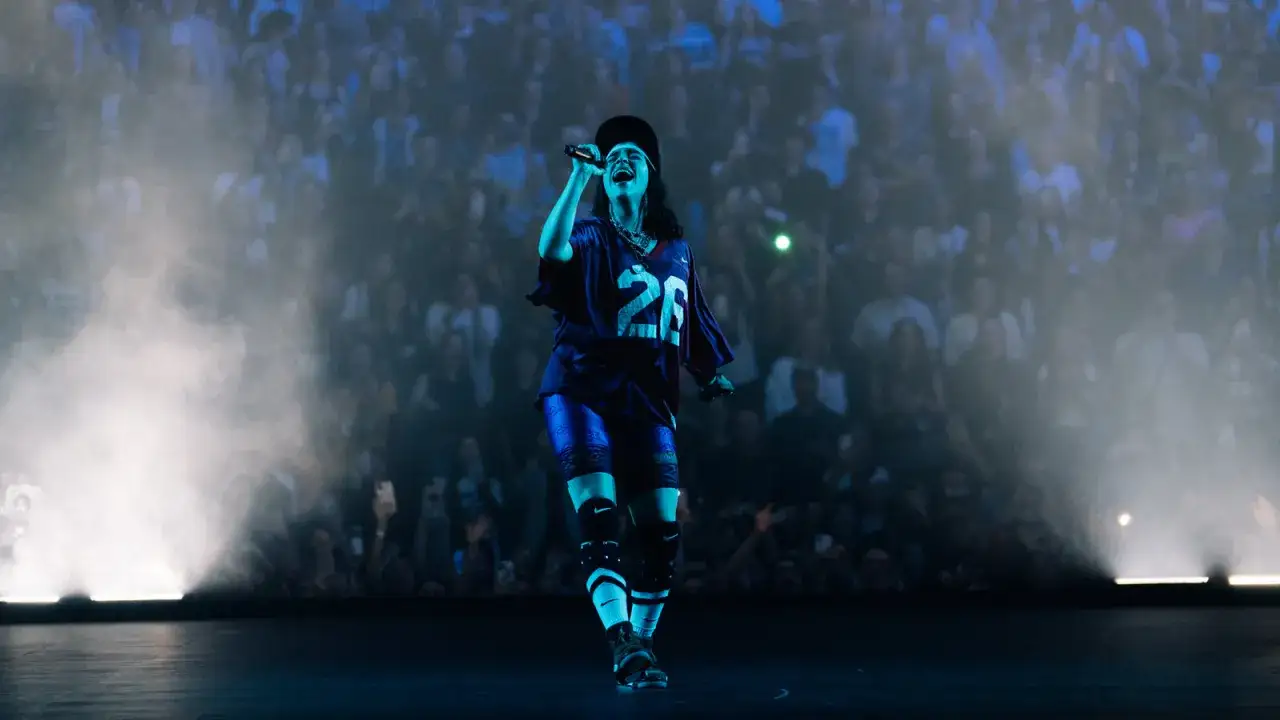 Billie Eilish teraz na scenie, w sportowej koszulce i z mikrofonem w dłoni, śpiewa dla tłumu fanów.