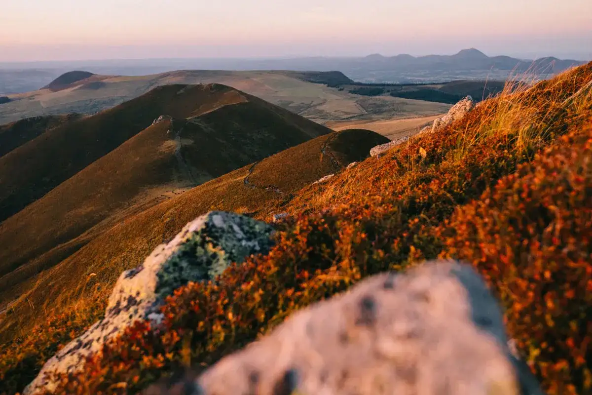 Vue panoramique des collines ondulantes et des volcans lointains, baignés par la lumière dorée du coucher de soleil. Un sentier serpente sur les pentes, invitant à explorer le tour des volcans d'Auvergne.