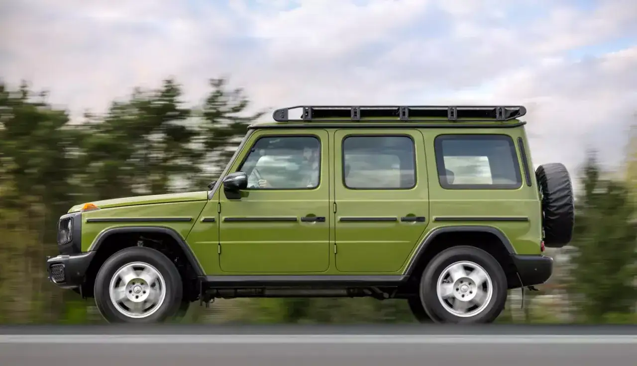 Zielony Mercedes-Benz Klasy G z bagażnikiem dachowym i kołem zapasowym na tylnej klapie, jadący po drodze wśród drzew.
